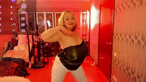 Media: Video of a plus-size blonde woman in a black leather corset, white pants, and red lipstick, posing confidently in a retro-themed bedroom with red walls, patterned wallpaper, and a plush carpet.