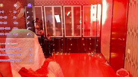 Media: A video of a dimly lit, red-hued room with a woman standing by a bed, wearing a black dress and red shoes. The background features a patterned wall and large windows with curtains.