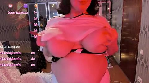 Media: Video of a woman with dark hair lifting her white top to reveal large breasts in a black bra, standing in a dimly lit room with a bed and colorful decor.