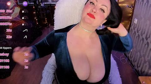 Media: Video of a curvy, fair-skinned woman with long black hair, wearing a deep V-neck velvet dress, seated in a dimly lit room. She has bold red lipstick and a pearl necklace.