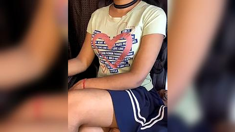 Media: Video of a young woman with light skin, wearing a white t-shirt with a heart graphic and navy blue shorts, sitting on a chair in a dimly lit room.