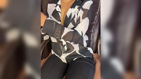 Media: Video of a person wearing a black-and-white patterned blouse and black pants, seated in a leather chair. The background is blurred.