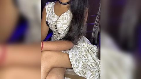 Media: Video of a young woman with long black hair, wearing a white, patterned dress with a high neckline, sitting on a chair in a dimly lit room.