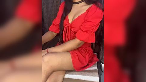 Media: Video of a woman in a red, short-sleeved, V-neck dress with a black choker, seated on a dark woven chair, blurred background.