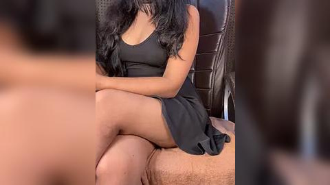 Media: Video of a dark-skinned woman with long, wavy black hair, wearing a short black dress, sitting on a beige couch, blurred background.