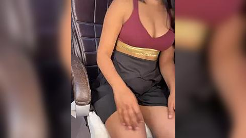 Media: Video of a woman with medium brown skin, wearing a burgundy sports bra and black shorts, seated on a massage chair with a leather backrest.