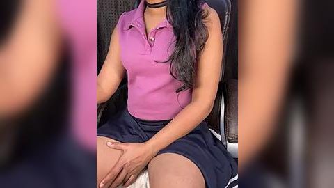 Media: Video of a woman with medium brown skin, wearing a pink polo shirt and dark blue skirt, sitting in a chair. Her long black hair flows over her shoulders.