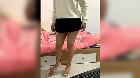 Media: Video of a light-skinned person wearing a cream sweater and black shorts, standing in a bedroom with a pink bedspread, white dresser, and keyboard on the floor.