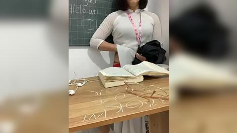 Media: Video of a woman with shoulder-length brown hair in a white blouse and black pants, holding a black bag, standing near a wooden desk with a book and a chalkboard behind her.