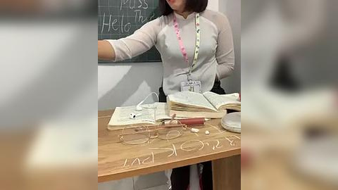 Media: Video of a woman in a white cardigan with a pink name tag, teaching at a wooden desk with open books, chalkboard, and \"Pssst Hello\" written on the desk.