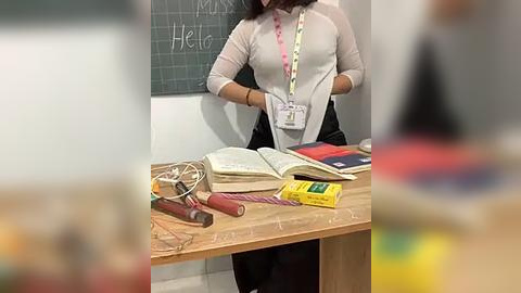 Media: Video of a woman in a white cardigan, black pants, and lanyard, standing behind a wooden desk with books, pens, and a lanyard. Chalkboard with \"Mrs. Heil\" in the background.