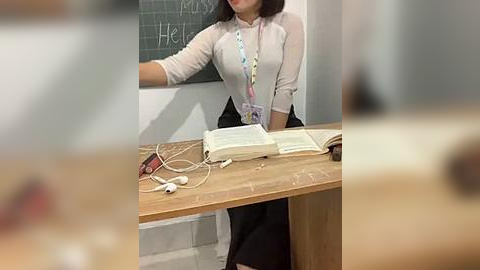 Media: Video of a woman in a semi-transparent white blouse, sitting at a wooden desk with an open book, a phone, and a lanyard. She's leaning against a chalkboard with \"Hello\" written on it, in a classroom setting.