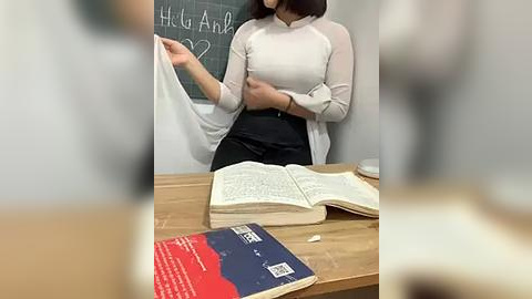 Media: A video of a woman with a medium build and short dark hair, wearing a white top and black skirt, teaching at a wooden desk with an open book and a red book.