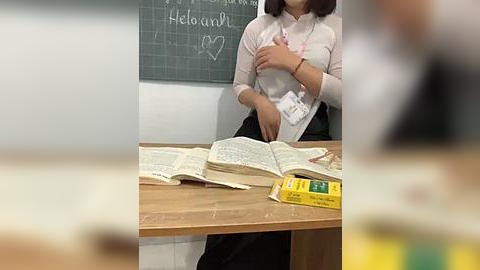 Media: Video of an Asian woman with shoulder-length black hair, wearing a white blouse and black skirt, seated at a wooden desk in a classroom. Books and a yellow box are on the desk. Chalkboard behind her.