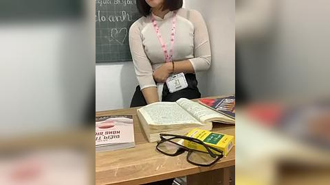 Media: Video of an Asian woman in a white blouse and black skirt, standing at a wooden desk with open books and reading glasses. A pink lanyard and badge hang around her neck.