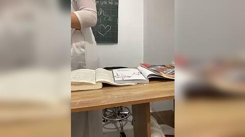 Media: Video of a person in a white lab coat standing at a wooden table with open books and a chalkboard in the background.