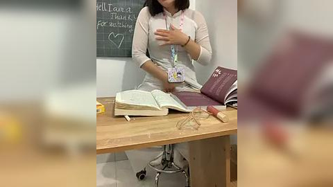 Media: Video of an Asian woman with short black hair, wearing a beige long-sleeve top, seated at a wooden desk, holding a book. A chalkboard behind her reads \"Welcome to the Library\" with a heart symbol.