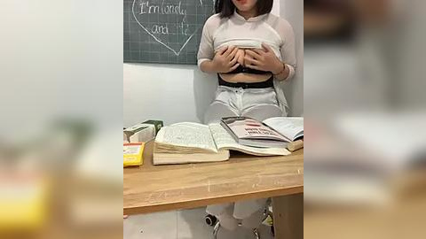 Media: Video of an Asian woman with medium build, wearing a sheer top, lifting it to reveal her breasts, sitting at a wooden desk with books and a green chalkboard in the background.