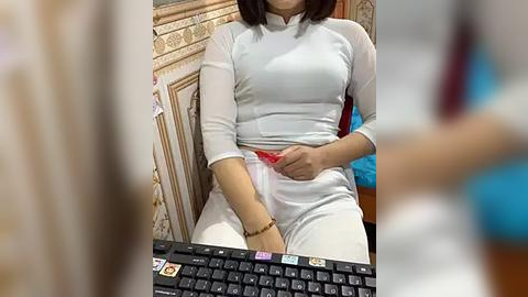 Media: Video of a woman in a white, long-sleeved top and white pants, seated in a room with beige, patterned wallpaper. She's adjusting her pants, revealing a red underwear peek. The background is blurred, showing a keyboard and part of a person in the foreground.