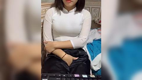 Media: Video of an Asian woman with medium-length black hair, wearing a white long-sleeve top and black pants, sitting on a bed with a blue blanket and white sheets. The background shows a beige wall and a decorative piece.