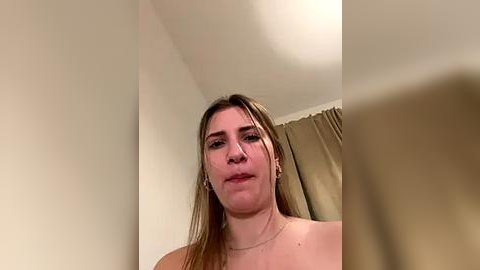 Media: Video of a young Caucasian woman with fair skin, straight brown hair, and a neutral expression, standing indoors against a beige wall and curtain backdrop.