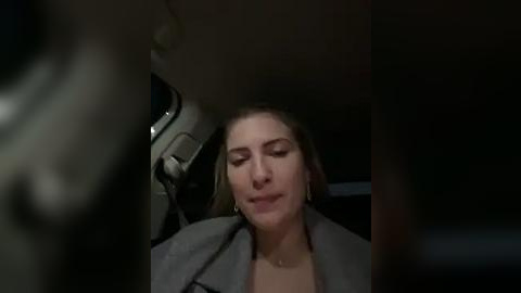 Media: Video of a woman with light skin, wearing a gray blazer, sitting in a car with dim lighting. She appears relaxed with her eyes closed, suggesting a moment of calm or sleep.