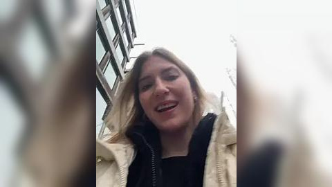 Media: A video of a woman with shoulder-length blonde hair, wearing a beige jacket and a black top, smiling widely while being videoed from a low angle. The background shows a city street with tall buildings and an overcast sky.