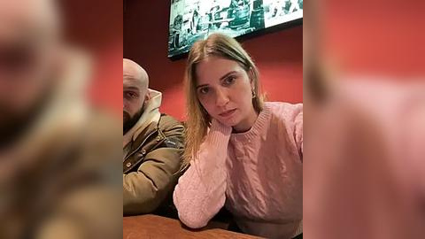 Media: Video of a young woman with straight blonde hair, wearing a pink sweater, sitting next to a bald man in a brown jacket, both looking at a TV screen showing a documentary.