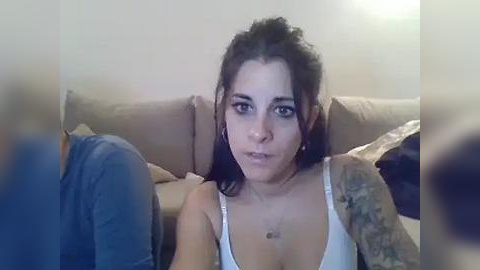 Media: A video of a young woman with dark hair and tattoos, wearing a white tank top, sitting on a beige couch in a dimly lit room.