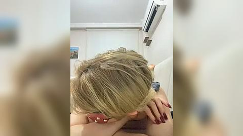 Media: Video of a blonde woman with a ponytail, lying on her stomach on a couch, with a light-colored wall and air conditioner in the background.