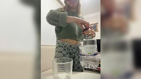 Media: Video of a woman with long blonde hair wearing a green fuzzy sweater and patterned pants pouring water into a clear plastic bottle. The background shows a cluttered kitchen counter with more bottles and a blurred, out-of-focus person.