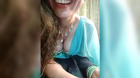 Media: Video of a smiling woman with long brown hair, wearing a turquoise blouse, multiple necklaces, and bracelets, partially cropped, with blurred background.