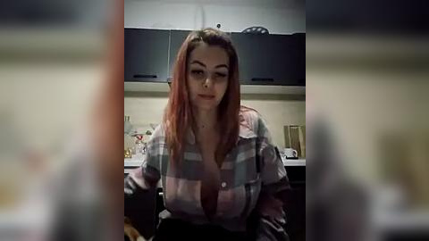 Media: A video of a Caucasian woman with long, straight auburn hair, wearing a plaid shirt, seated in a modern kitchen. The background features dark cabinets, a countertop with utensils, and a blurred view of the room.