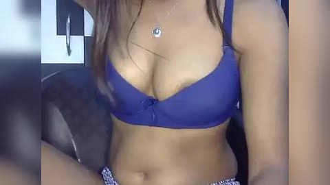 Media: A video of a woman with medium brown skin wearing a blue lace bra and patterned shorts, sitting on a dark couch.