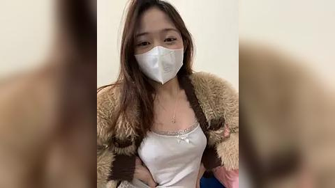 Media: Video of an Asian woman with long brown hair, wearing a white mask, white tank top, and brown faux fur jacket, indoors with blurred background.