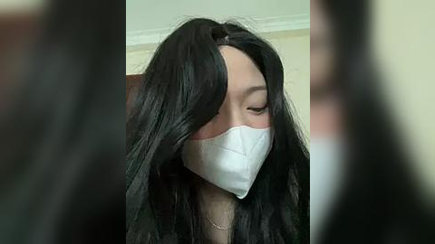 Media: Video of a woman with long, black hair wearing a white surgical mask, seated indoors, blurred background.