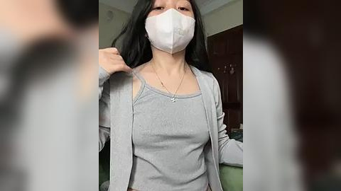 Media: Video of a woman with long black hair, wearing a white face mask, grey top, and cardigan, adjusting her mask in a domestic setting with wooden cabinets and green carpeted floor.