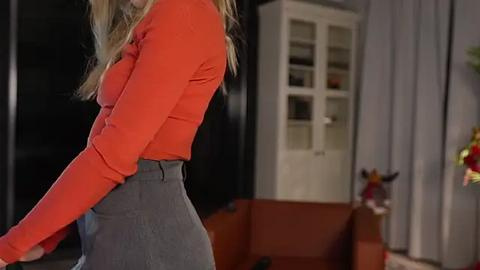 Media: Video of a blonde woman in a red long-sleeve top and high-waisted gray skirt, standing in a modern living room with a white cabinet and a brown sofa, dim lighting.