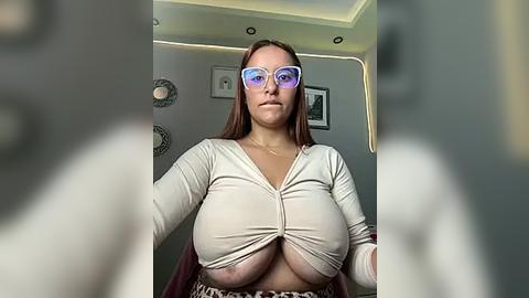 Media: Video of a fair-skinned woman with large breasts, wearing a tight beige top and purple reflective glasses, in a dimly lit room with white walls and abstract art.