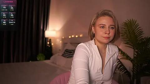 Media: Video of a blonde woman with a short bob, wearing a white blouse, sitting on a bed in a cozy, dimly lit room.