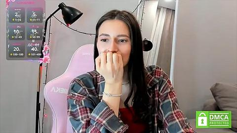 Media: Video of a young woman with long dark hair, wearing a plaid shirt, covering her mouth, seated in a pink gaming chair.