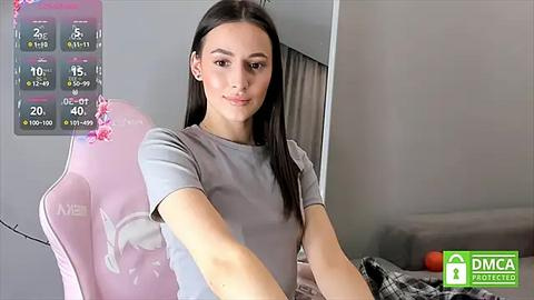 Media: Video of a young Caucasian woman with straight black hair, wearing a gray T-shirt, seated in a pink gaming chair in a modern, minimalistic room with a digital temperature display.