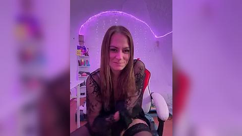 Media: A video of a young Caucasian woman with straight, light brown hair, wearing black lace lingerie, seated on a gaming chair in a dimly lit, purple-lit room with shelves and a desk in the background.