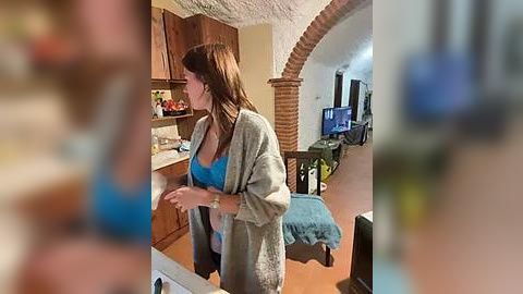 Media: A video of a woman in a blue bra and gray cardigan, standing in a cozy, warmly lit kitchen with wooden cabinets and a countertop.