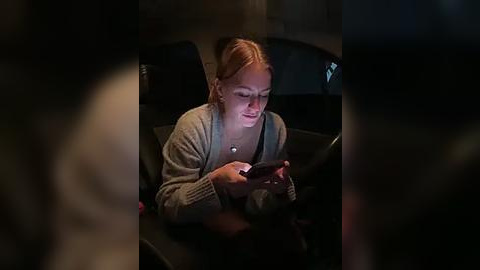 Media: Video of a young woman with blonde hair, wearing a grey cardigan, deeply engrossed in her smartphone, sitting in a dimly lit, dark interior, possibly a car or bus.