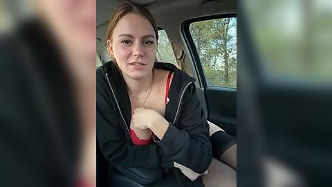 Media: Video of a young woman with fair skin, brown hair, and a red bra strap showing under her black hoodie, sitting in a car, blurry background.