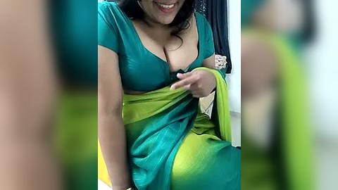 Media: Video of a South Asian woman with medium skin tone, wearing a teal and lime green sari, partially revealing cleavage, smiling, indoors.