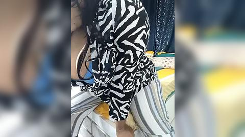 Media: Video of a person with long black hair, wearing a black and white patterned top, exposing a blue bra, and striped pants, sitting on a bed with yellow and green pillows.