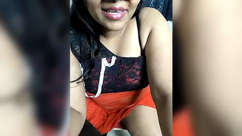 Media: Video of a young Asian woman with long black hair, wearing a black lace top and bright orange shorts, smiling with light pink lipstick.