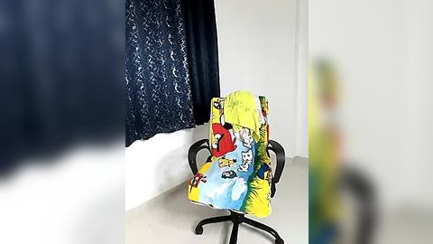 Media: Video of a colorful, cartoon-printed chair in a minimalistic room with white walls and a navy-blue curtain. The chair has a yellow, blue, and red pattern.